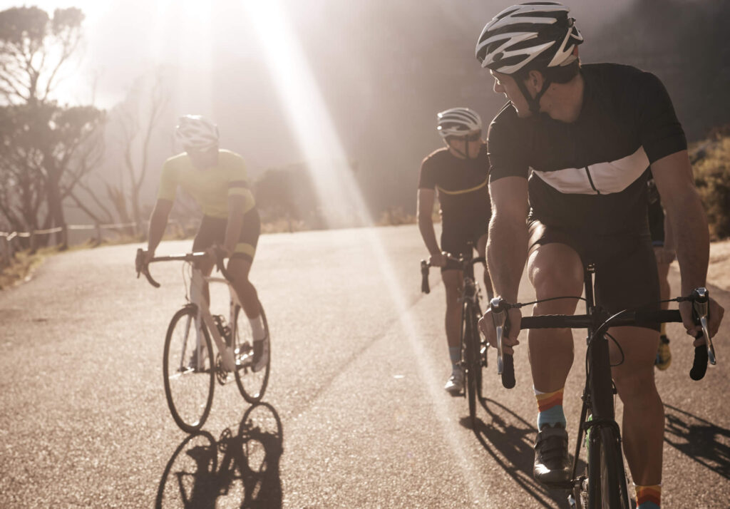 Tre cykelryttere i sollys cykler op ad en vej omgivet af natur. Rytterne bærer hjelme og cykeltøj, og billedet illustrerer stemningen i et cykelprogram.