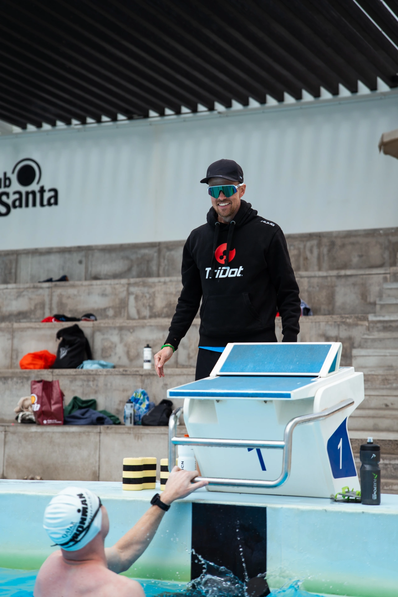 ironman-traener-coach-lykke-endurance Coach Lykke Endurance Lifeguard in a black hoodie and cap stands near a blue-and-white lifeguard chair beside a swimming pool, watching the swimmers behind him