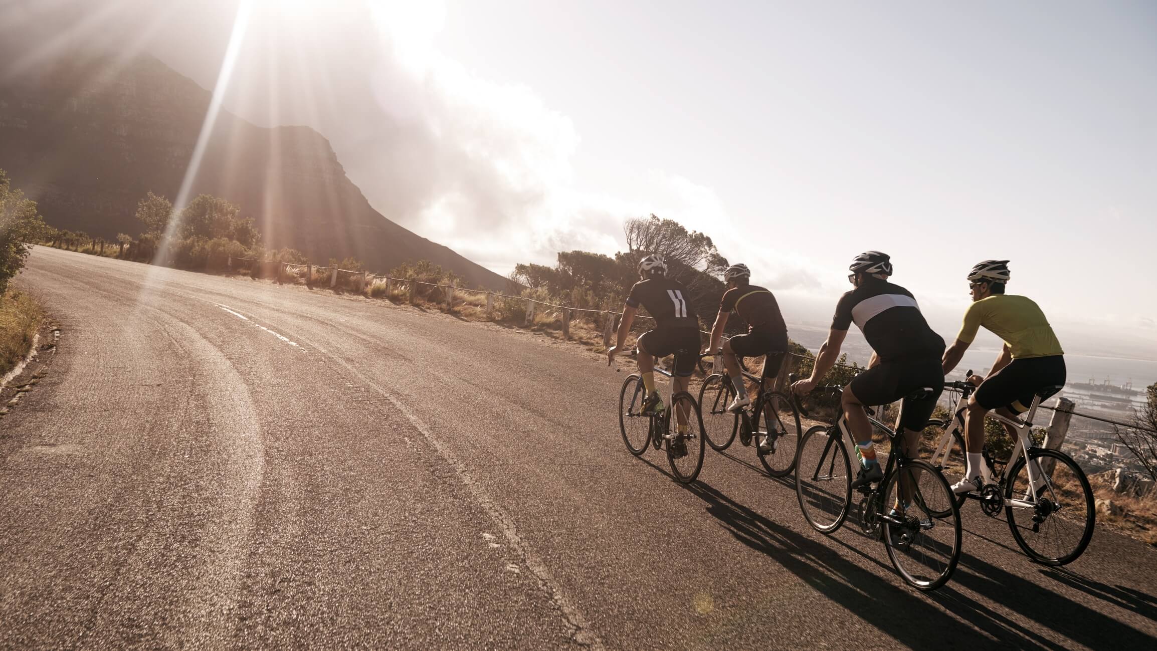 Fire cyklister kører op ad en solbeskinnet landevej med udsigt over havet og byen i baggrunden. Til venstre ses en introduktion til personlige træningsprogrammer i triatlon, cykling, gravel og løb.