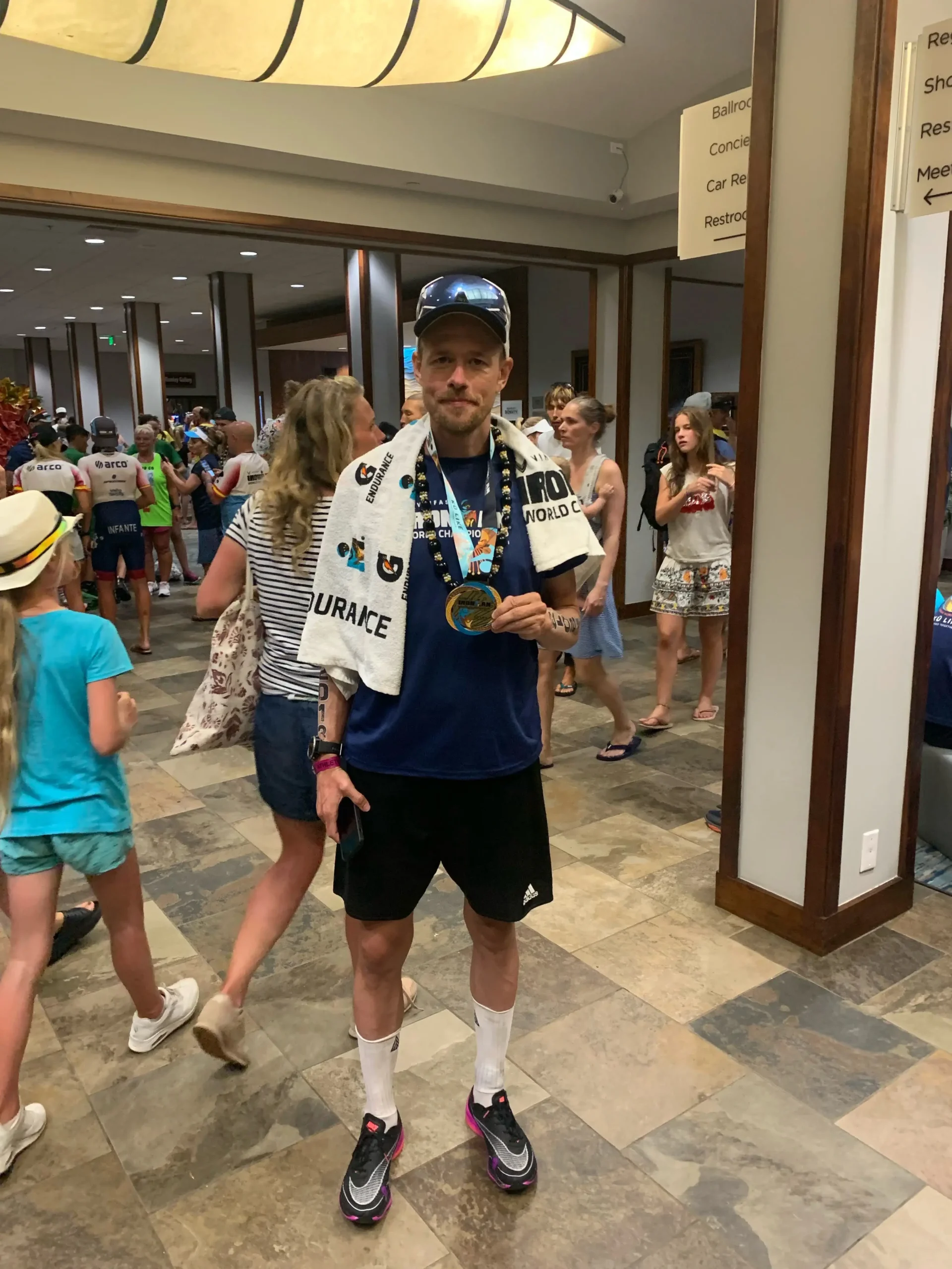 thomas-paa-hawaii Coach Lykke Endurance | Triatlon • Cykling • Løb Man with medal in gym lobby, wearing towel around shoulders, cap with sunglasses, blue shirt and black shorts.