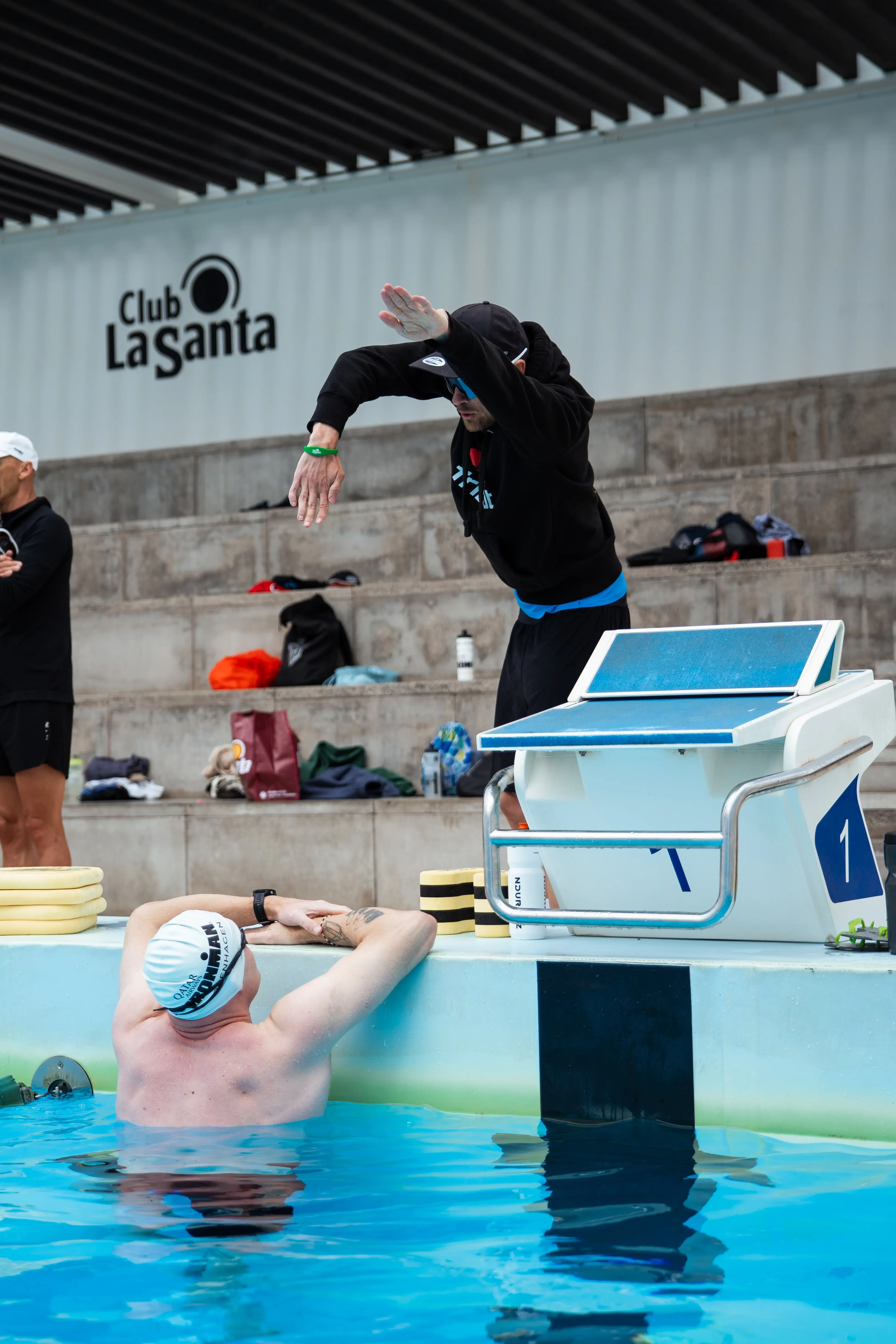 traener-thomas-bassin-1 Coach Lykke Endurance A swimmer wearing a cap and goggles stands on a diving block, preparing to jump into a bright blue pool with a “Club La Santa” wall in the background.